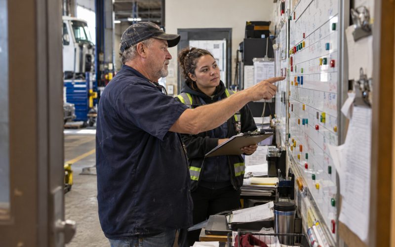 Two people at a maintenance planning board in a shop office, one pointing at a card